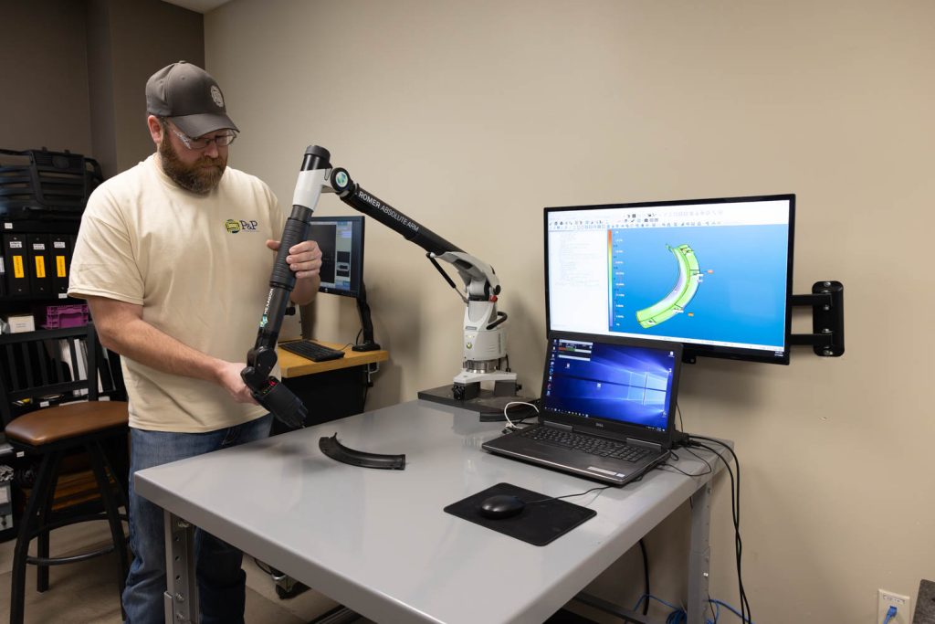 A man uses a portable 3D measuring arm to scan a plastic part on a table. A computer displays 3D modeling software with the scanned object, likely for injection molding. The workspace contains various equipment and office furniture.