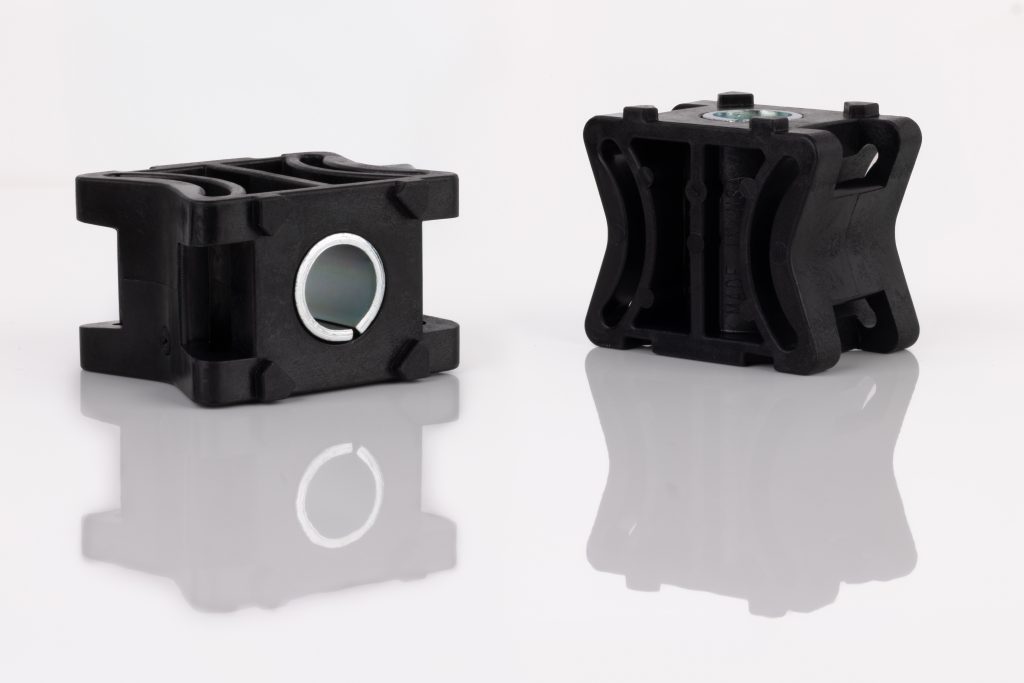 Two black injection molded plastic parts with circular metal inserts are shown on a reflective white surface. One part faces forward displaying the insert; the other is angled, revealing side and top details.